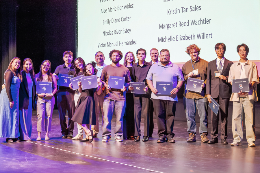 students stand on stage holding achievement awards during a recognition ceremony with names projected on a screen behind them