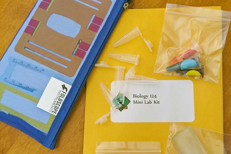 biology 124 mini lab kit with a foldscope microscope, pipettes, test tubes and other experiment supplies arranged on a table
