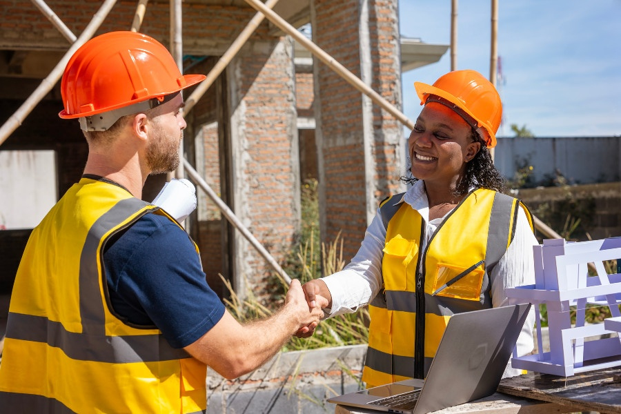 two construction professionals wearing hard hats and safety vests shake hands beside building plans, a laptop and a construction model at a job site