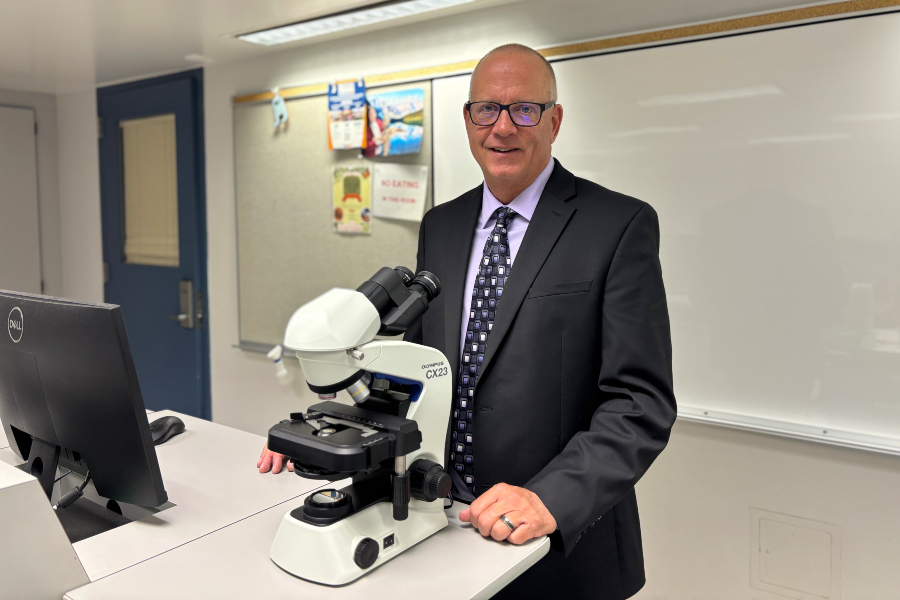 Dr. Eric Rabitoy stands beside a microscope in a science classroom with a computer, whiteboard and classroom materials in the background