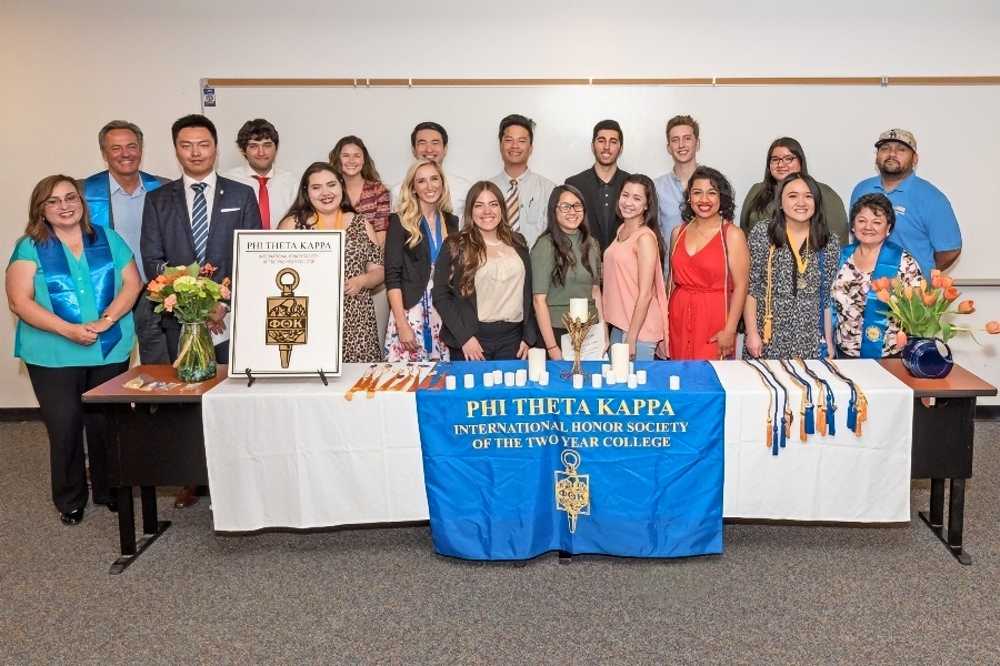 citrus college phi theta kappa members and advisors pose behind a table displaying the honor society banner medals flowers and ceremony items