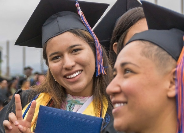 graduate in cap and gown holding a diploma cover and making a peace sign during commencement ceremony while fellow graduates gather nearby