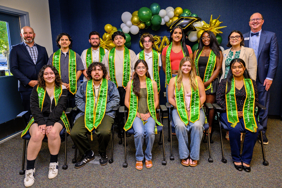 dual enrollment graduates wearing green stoles pose with college representatives at a recognition ceremony decorated with green, gold and white balloons