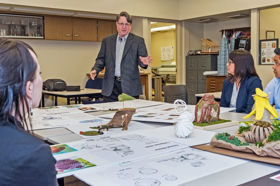architecture instructor speaks with students gathered around a classroom table covered with architectural drawings, sketches and three-dimensional design models