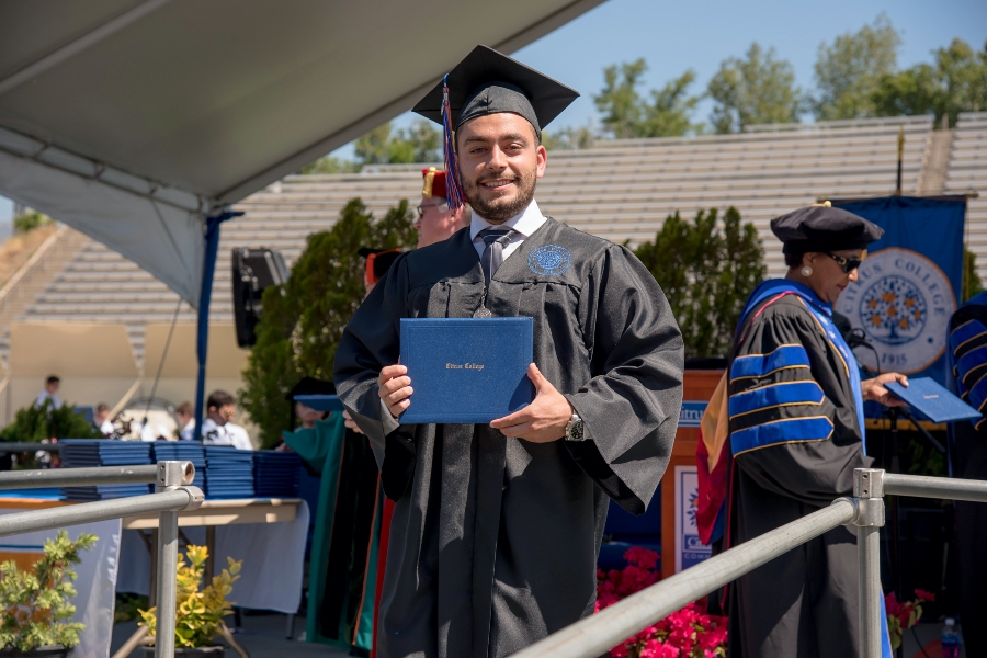 johnny elie baradii smiles in a black cap and gown while holding a blue citrus college diploma cover, with dr. barbara r. dickerson in academic regalia in the background