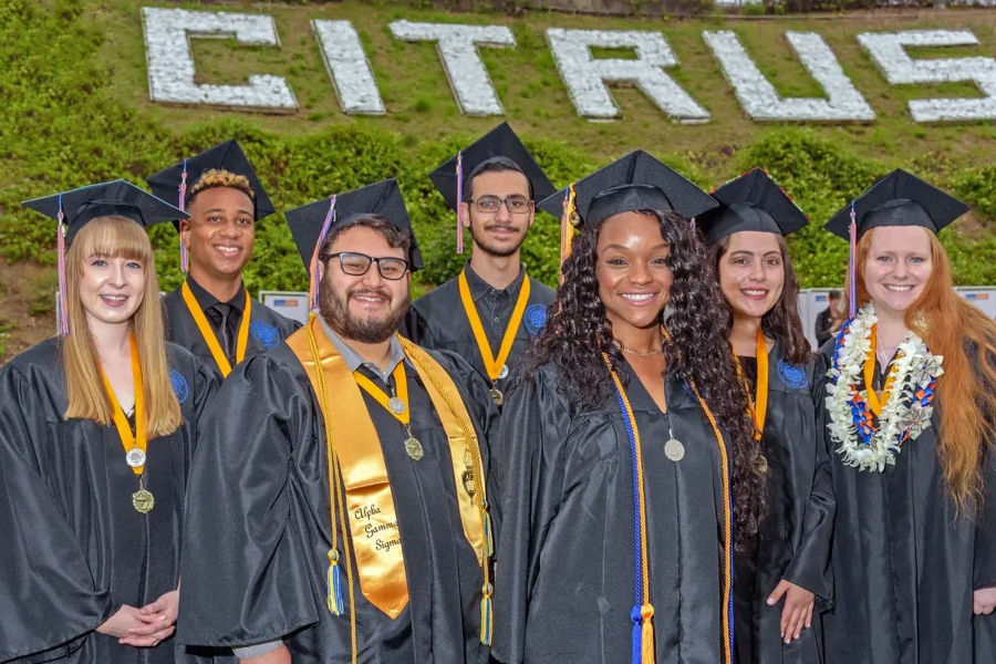 seven citrus college graduates in caps and gowns pose together in front of the citrus hillside sign