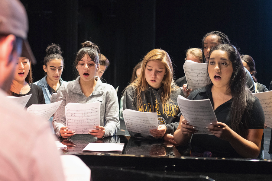 students rehearse a choral performance around a piano as an instructor accompanies them