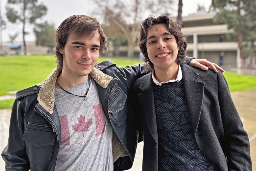 two citrus college students stand together outdoors on campus, one wearing a black leather jacket over a gray canada t-shirt and the other wearing a dark coat and patterned sweater with his arm around his classmate's shoulder