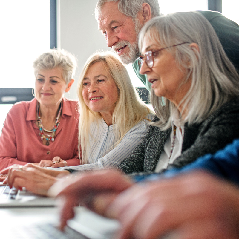 group of older adults gathered around a laptop, smiling and engaged as they learn together in a bright, collaborative setting