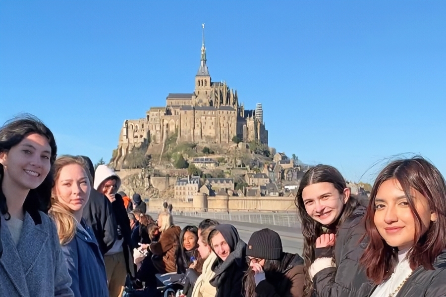 students pose in front of mont-saint-michel in normandy, france, with the historic abbey and fortified village rising behind them under a clear blue sky