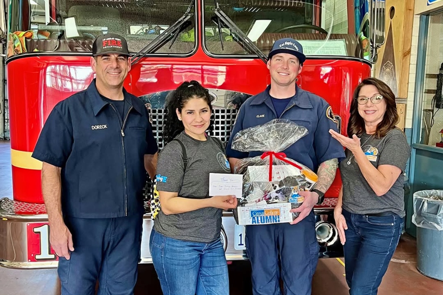 two firefighters and two women stand in front of a red fire engine inside a station; one firefighter holds a citrus college alumni gift basket while one woman holds a thank-you card and the other gestures toward him