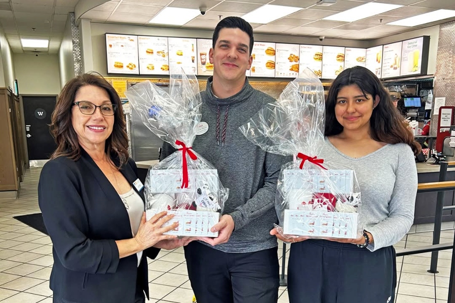 three people stand inside a chick-fil-a restaurant holding two wrapped gift baskets prepared for a citrus college alumni and friends firefighter appreciation visit