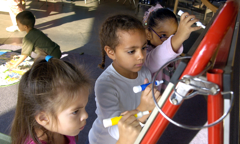 three early elementary girls intently creating art on an ease, while a little boy plays behind them