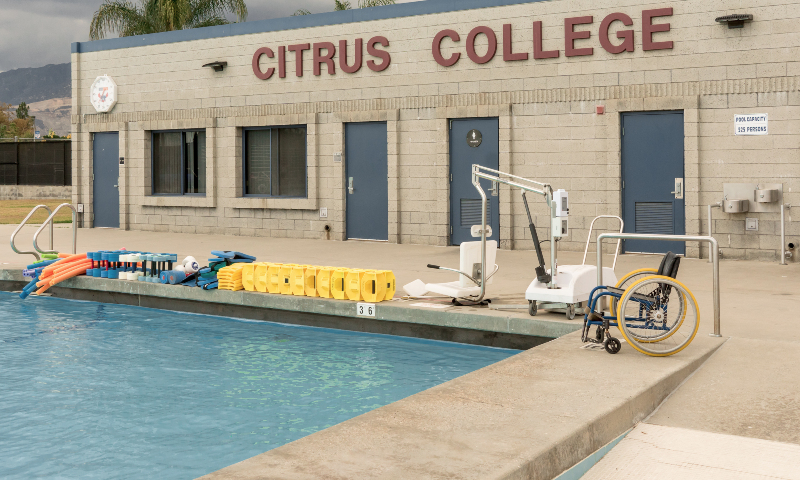 view of equipment used in the aquatics class next to the pool with a wheelchair near the pool ramp, and the aquatics center building in the background