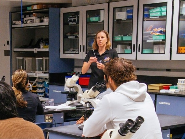 instructor speaks to students seated at microscopes during a laboratory class