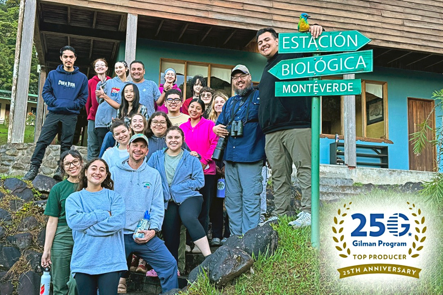 group of citrus college study abroad students and faculty standing outside a building at estación biológica monteverde in costa rica, gathered beside a green sign marking the biological station. gilman program 25th anniversary top producer badge appears in the lower corner