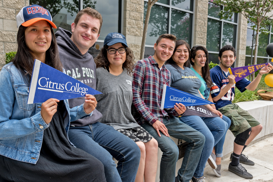 college students sit outside holding pennants and wearing apparel from transfer universities