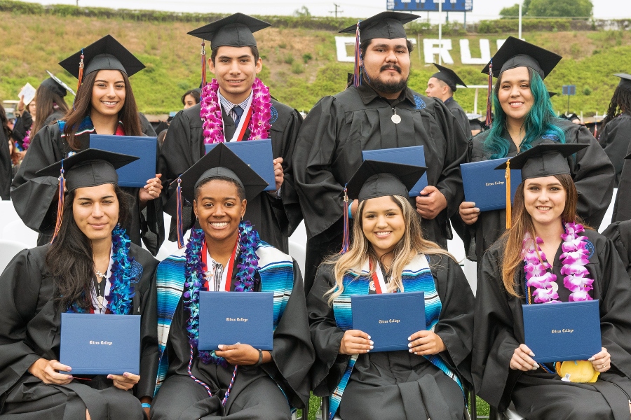 eight citrus college graduates in caps and gowns pose together holding diploma covers at an outdoor commencement ceremony