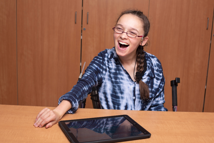 elina hughes laughs while seated behind an ipad she uses for assistive communication