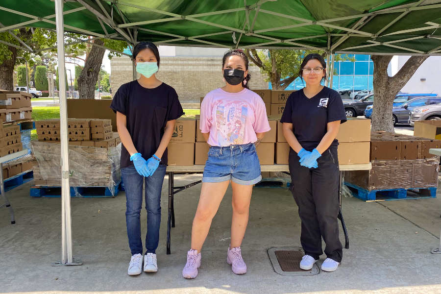 three volunteers wearing masks and gloves stand under a canopy in front of boxes at a campus food pantry event