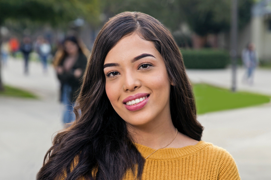 a smiling citrus college student poses outdoors in a mustard sweater with campus walkways softly blurred behind her