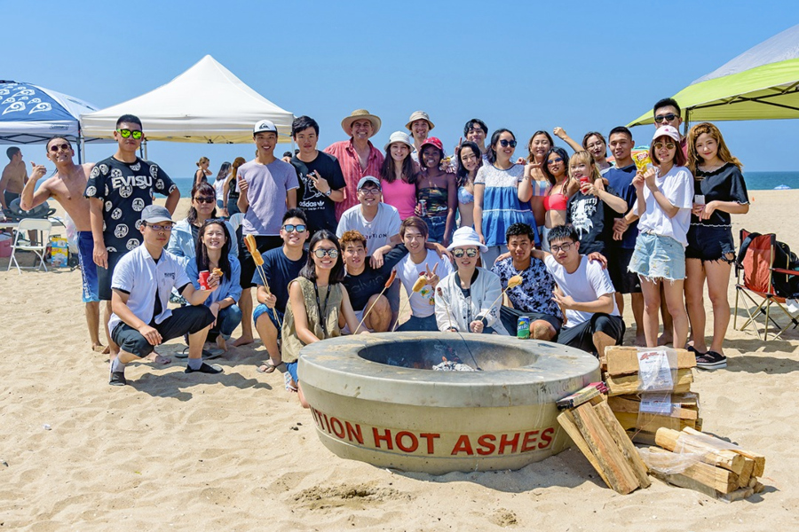 international students gather for a beach outing around a fire pit with tents and ocean in the background