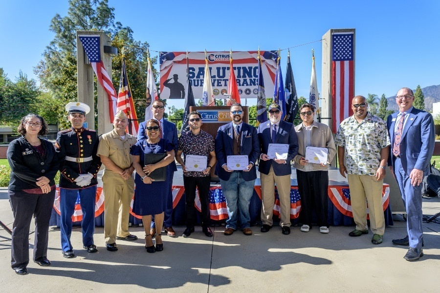saluting our veterans honorees and campus representatives pose in front of a patriotic stage with flags during an outdoor recognition event