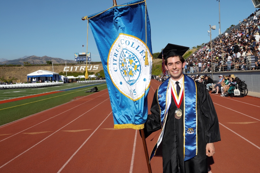student trustee aj hernandez in cap and gown holds a citrus college banner on the stadium track during commencement, with medals around his neck and graduates, guests and stadium commencement set up in the background