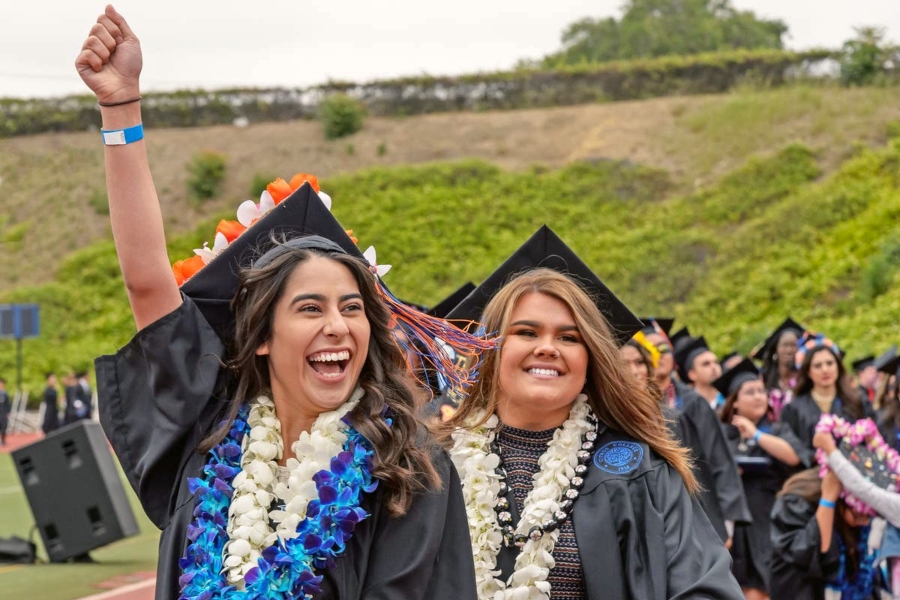 two citrus college graduates in caps and gowns wear flower leis as one raises her fist in celebration during commencement