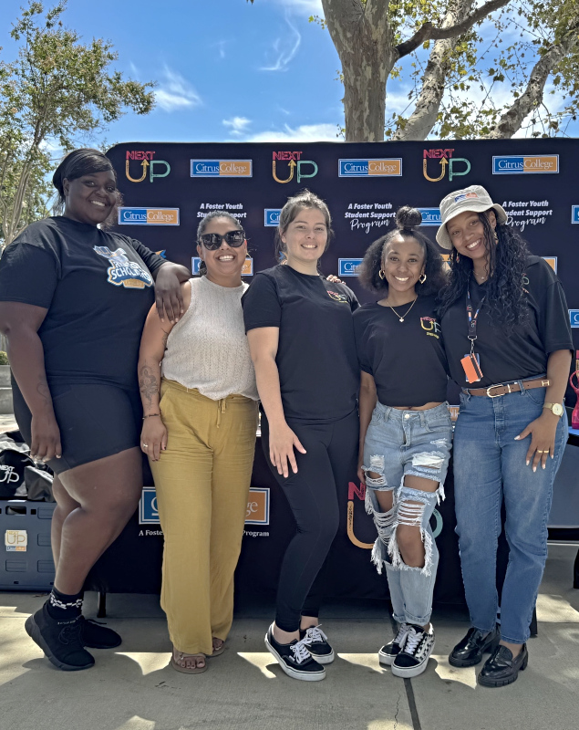 group of five smiling students and staff standing outdoors in front of a citrus college nextup step-and-repeat banner during a campus event, wearing casual clothing and posing together under trees on a sunny day
