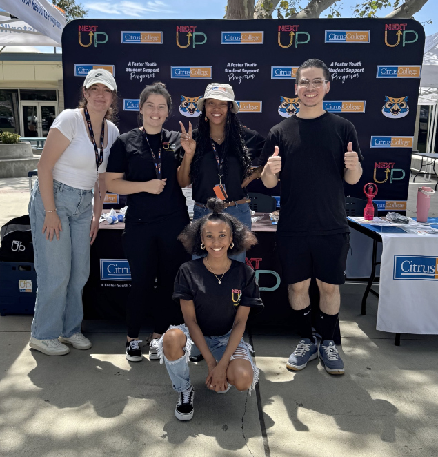 five students and staff posing at a citrus college nextup outreach table outdoors, smiling and giving thumbs up and a peace sign in front of a nextup and citrus college branded backdrop during a sunny campus event