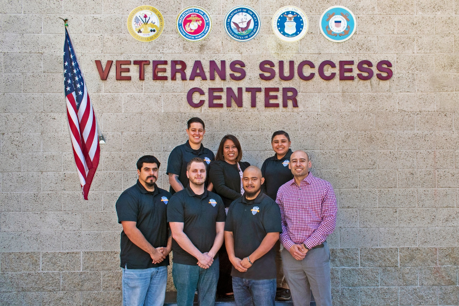 seven people pose beneath veterans success center signage, military branch emblems and an american flag