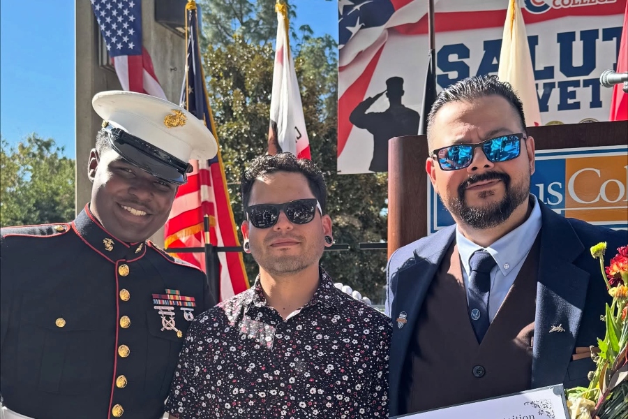 a u.s. marine in dress uniform stands with two men during citrus college's saluting our veterans ceremony, with american and california flags and the event banner displayed behind them