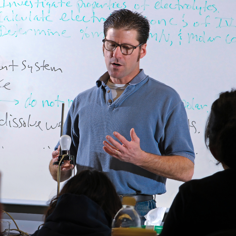 chemistry faculty, Terry Miles, presents to his class with a whiteboard behind him