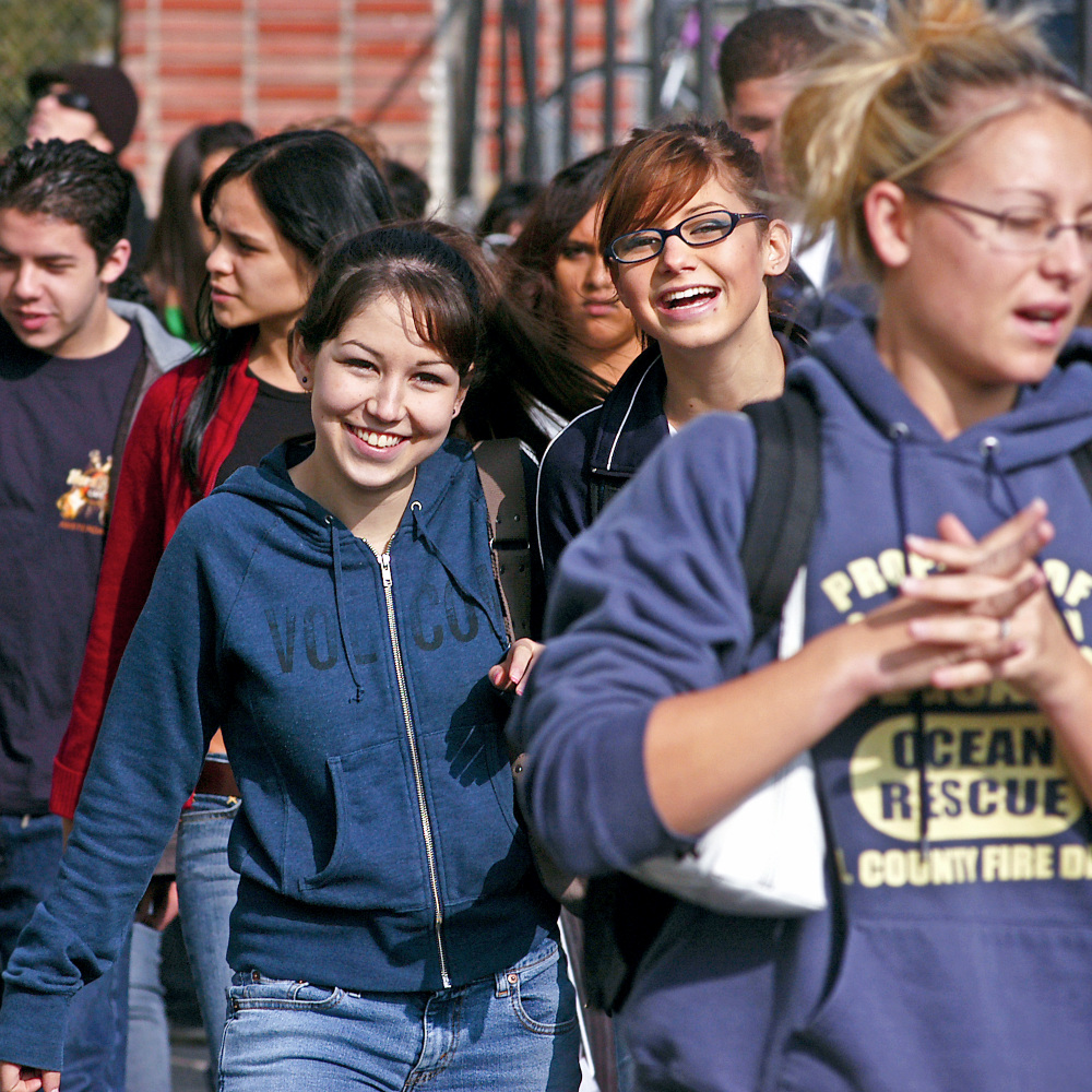 high school students walk together across campus, with two smiling young women near the center of the crowd