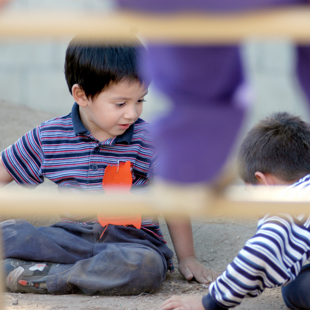 two young children play together outdoors, viewed through a wooden railing, with one child seated and holding an orange craft project