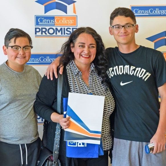 a woman holding citrus college promise materials stands between two smiling young men in front of a citrus college promise backdrop