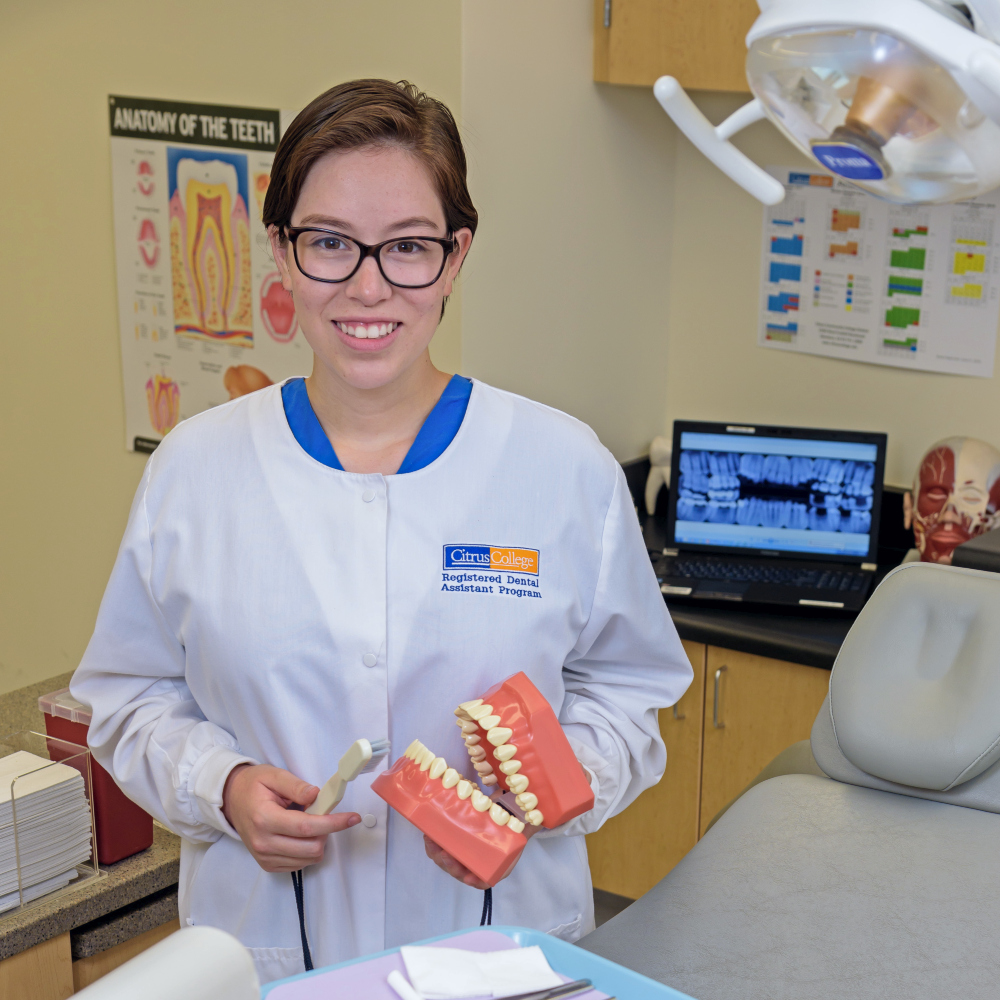 dental assisting student holds a toothbrush and model teeth in a campus dental lab with an exam chair, x-ray image and dental equipment in the background