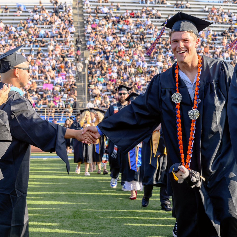 smiling graduate wearing a cap, gown, honor cords and medals shakes hands with another graduate during commencement as spectators fill the stadium bleachers