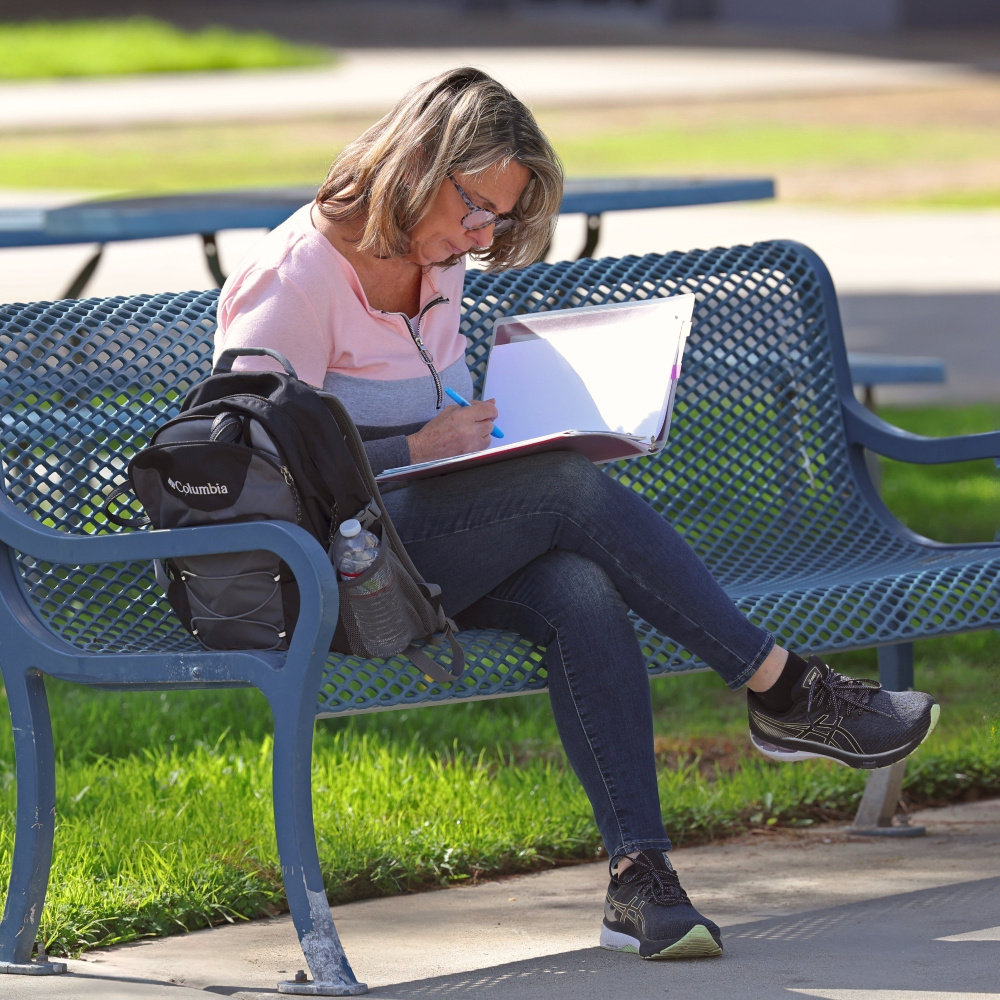an adult student writes in a binder while seated on a campus bench outdoors