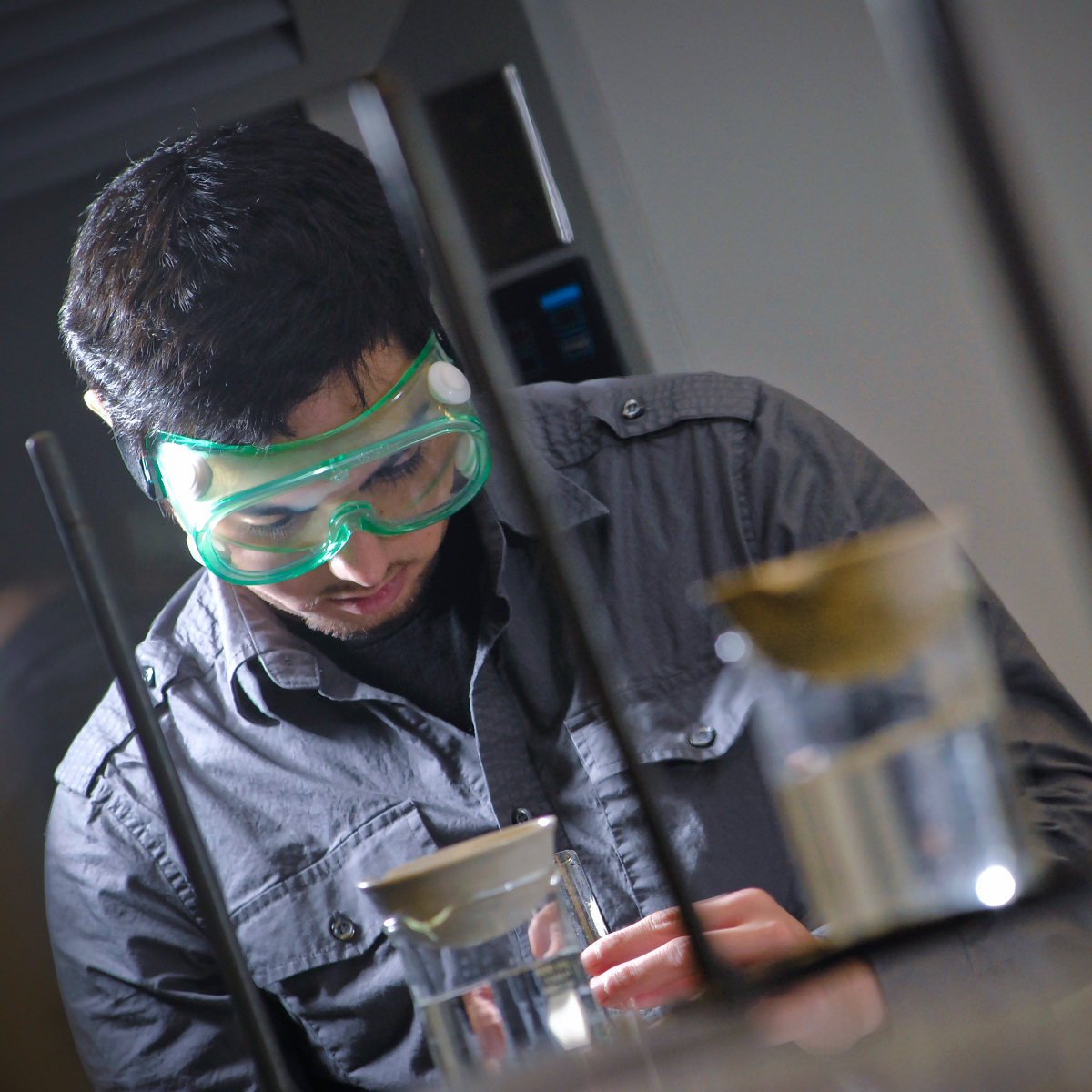 a student wearing green safety goggles examines laboratory glassware filled with clear liquid during a science experiment