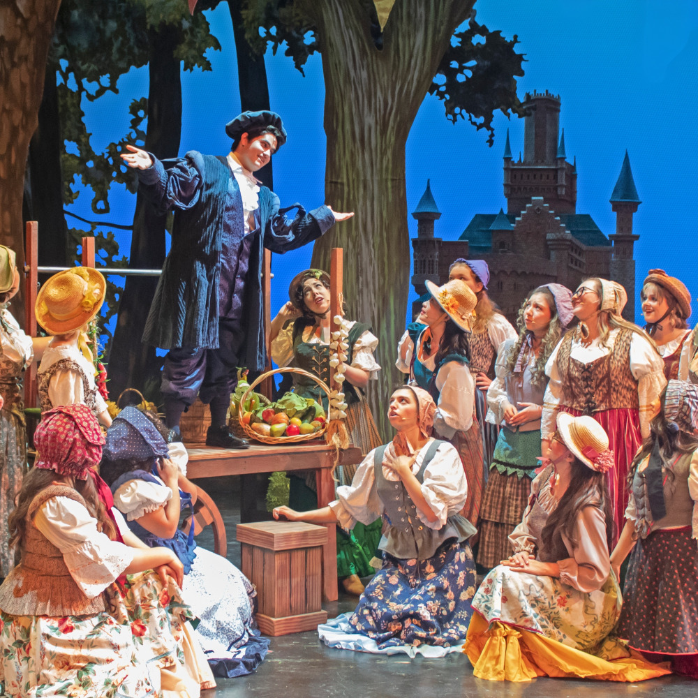 a performer in a blue period costume gestures from a raised platform as an ensemble in colorful rustic clothing gathers around on a theatrical set with trees and a castle backdrop