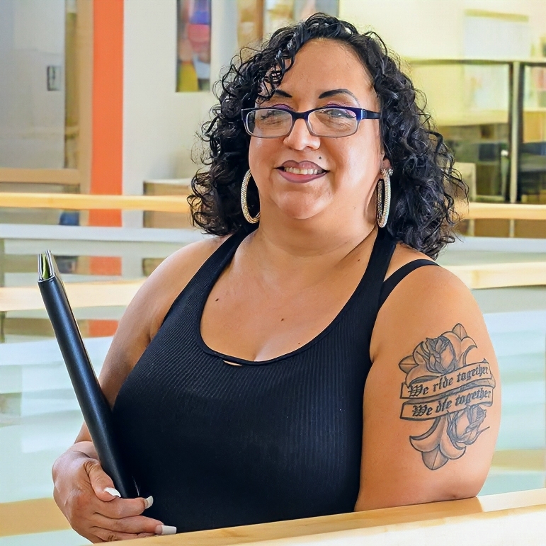 a woman with curly dark hair and glasses smiles while seated indoors, wearing a black sleeveless top and hoop earrings and holding a black diploma cover; a floral tattoo is visible on her upper arm