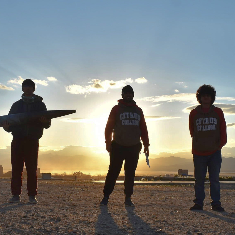 three citrus college students stand outdoors at sunset, with one student holding a model rocket