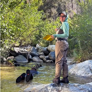 Emily Correia-Lambton standing on a rock in a shallow stream wearing chest waders and holding a yellow field notebook while conducting outdoor environmental fieldwork in a wooded area